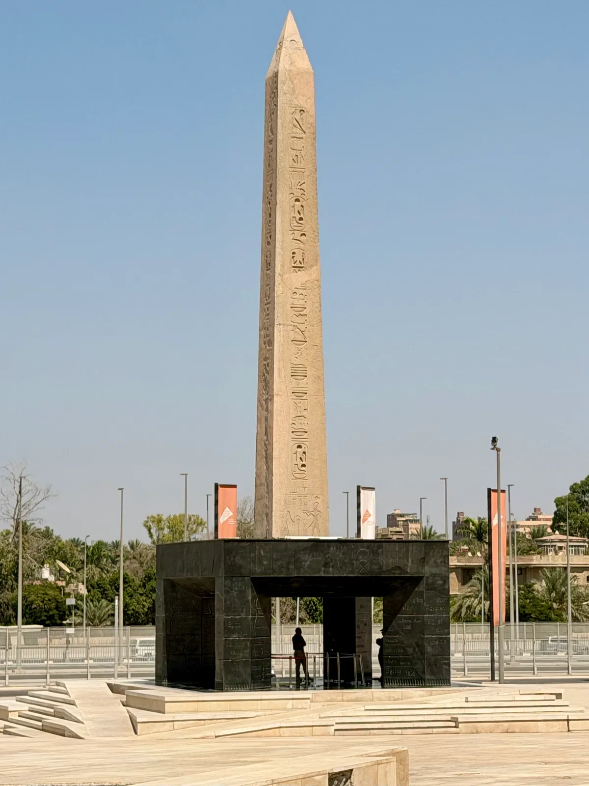 A tall red granite obelisk covered in hieroglyphs standing on a dark open-sided plinth in a paved plaza, with a visitor standing underneath it looking up into the base