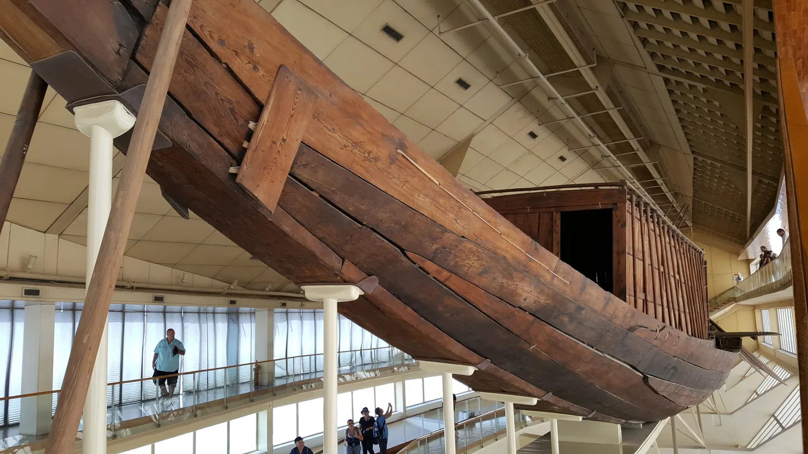 The hull of the Khufu ship, a long boat built from broad cedar planks curving up to a high prow, raised on supports inside a museum hall with visitors on a walkway below it