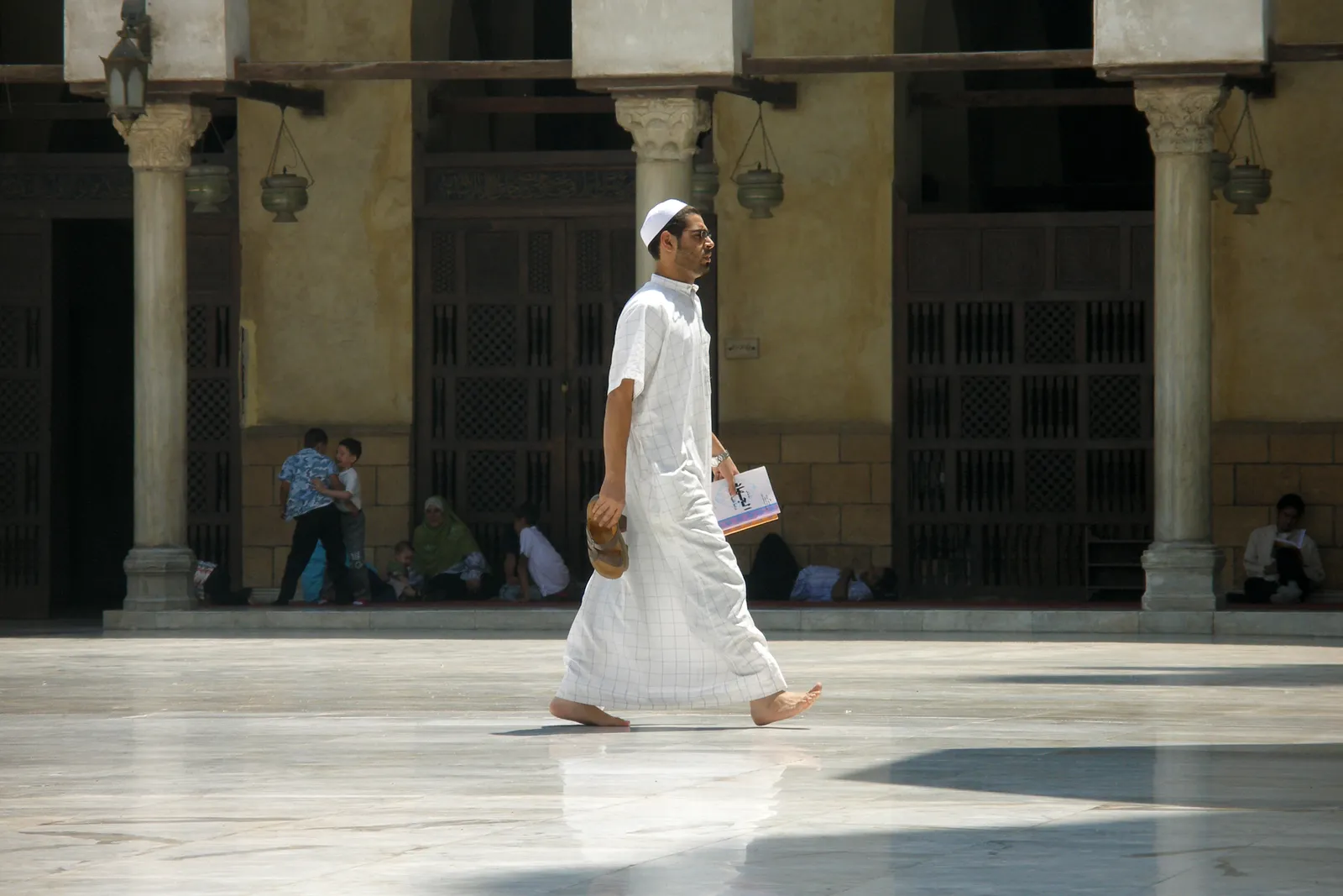 The open courtyard of Al-Azhar Mosque in Cairo, a man in white crossing the sunlit marble paving with an arcade of columns and hanging lamps in shadow behind him