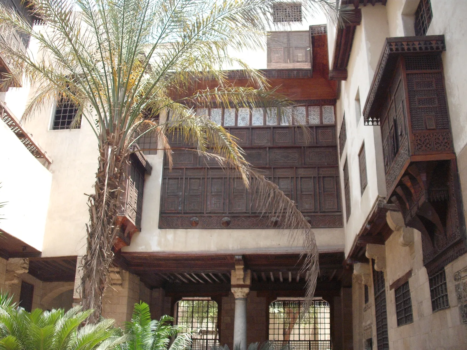 The inner courtyard of Bayt al-Suhaymi in Cairo, with dark latticed wooden window screens projecting from the upper floors over a stone-paved yard and a date palm growing in one corner