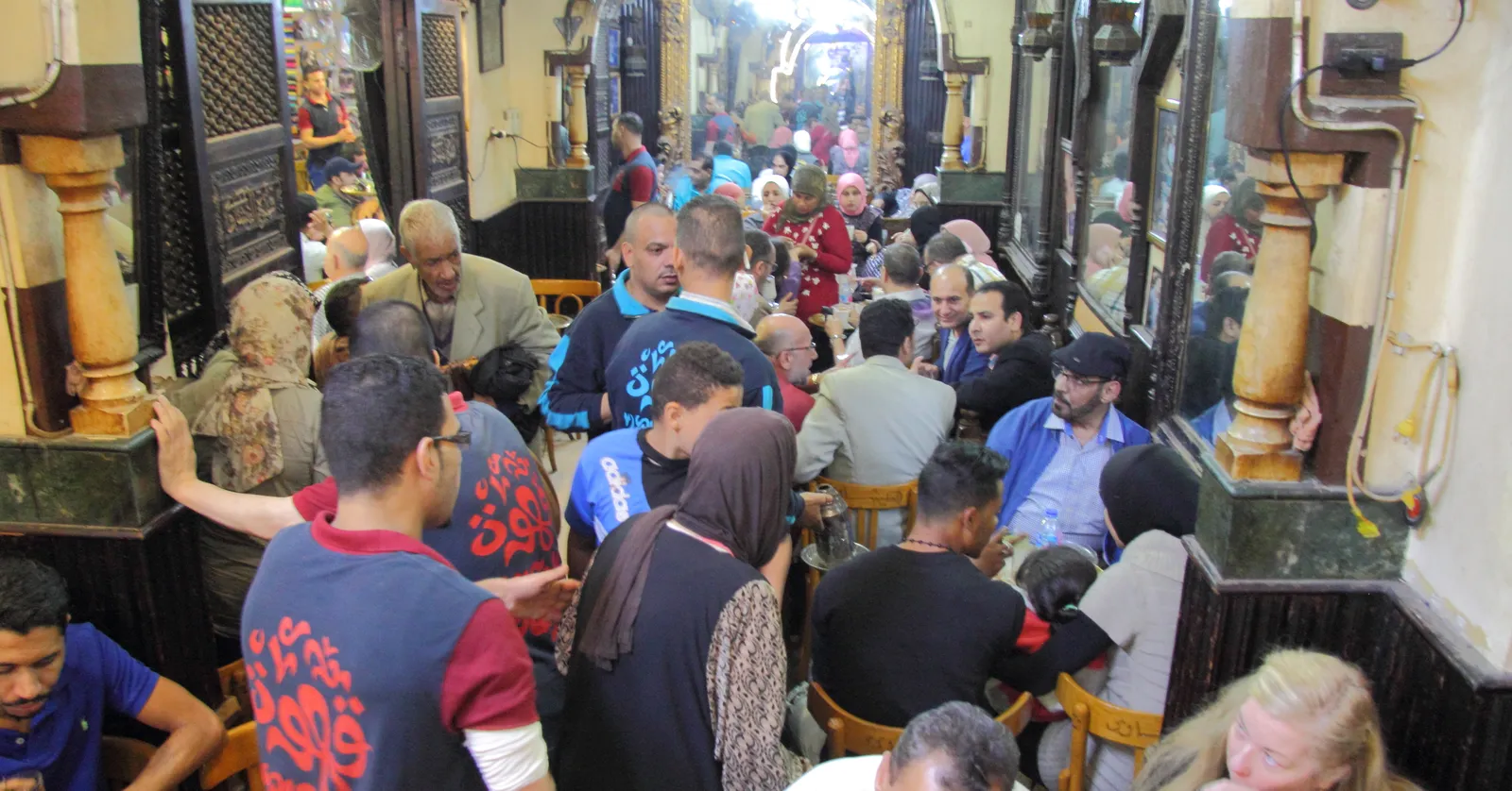 The packed alley of El Fishawi cafe in Khan el-Khalili, tall gilt-framed mirrors and carved wooden screens lining both walls above small tables with customers crowded between them