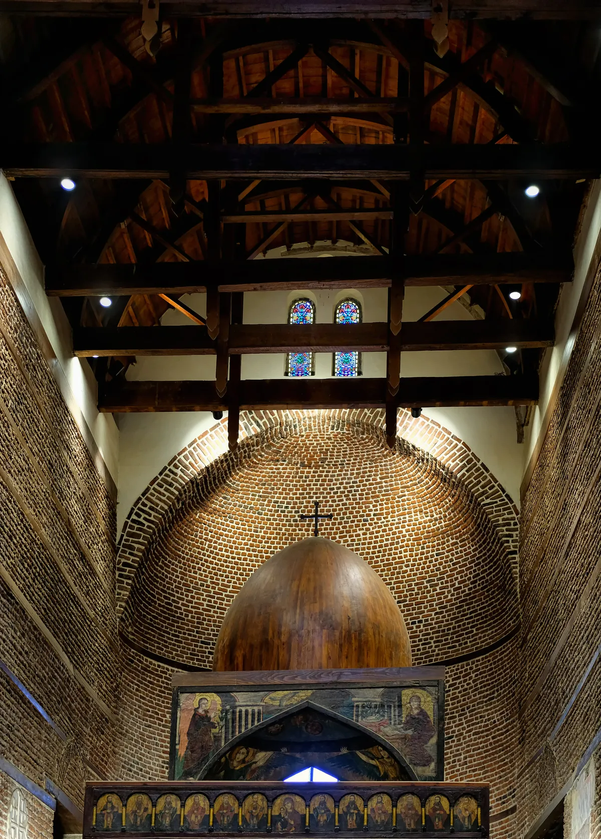 The interior of Abu Serga church in Old Cairo, a high open timber roof over brick walls with a brick half-dome above the altar screen and small stained-glass windows in the gable