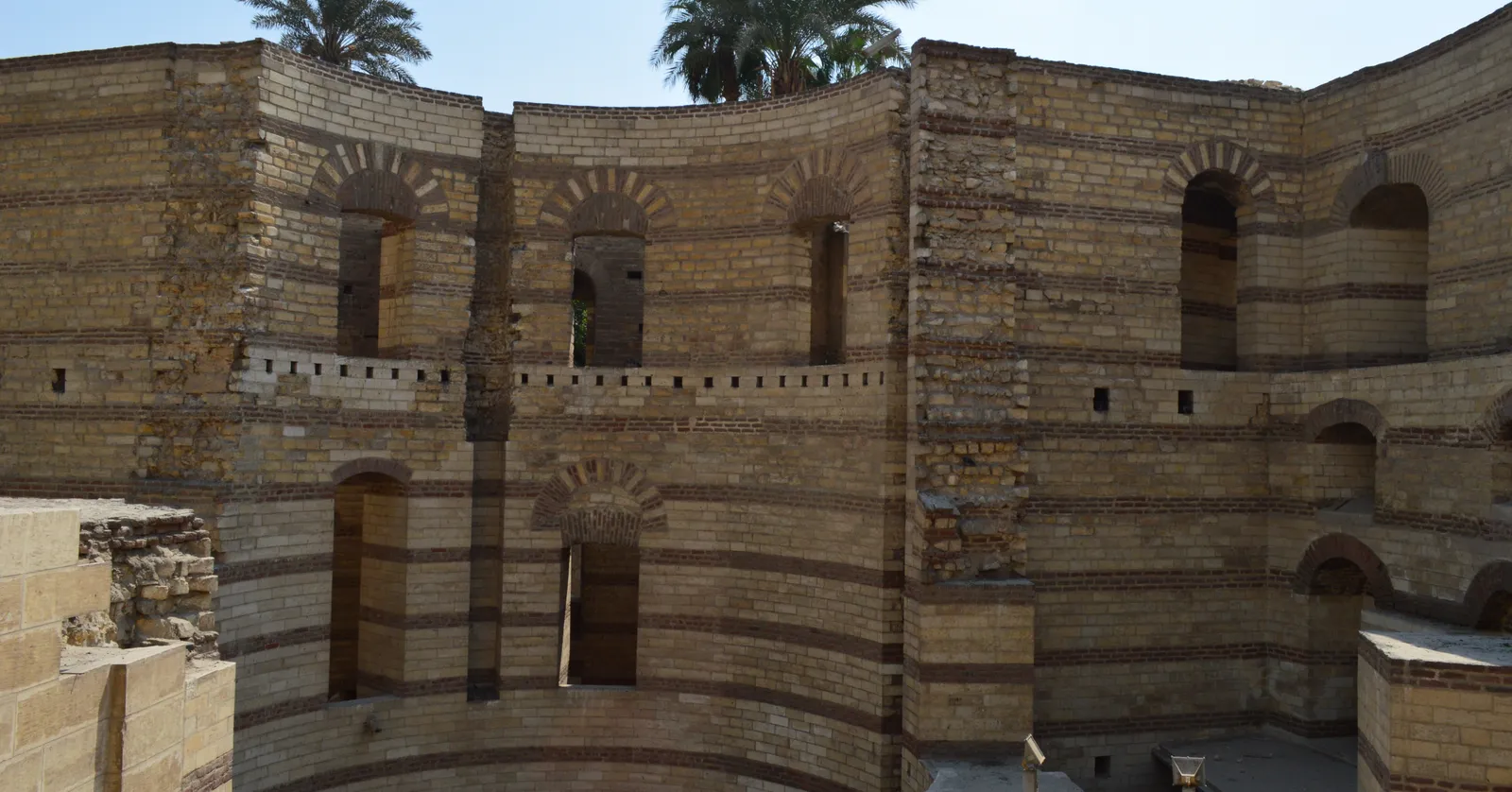 The curved brick and stone walls of a Roman tower of the Babylon fortress in Old Cairo, built in alternating bands with arched recesses set into the face and palms above the parapet
