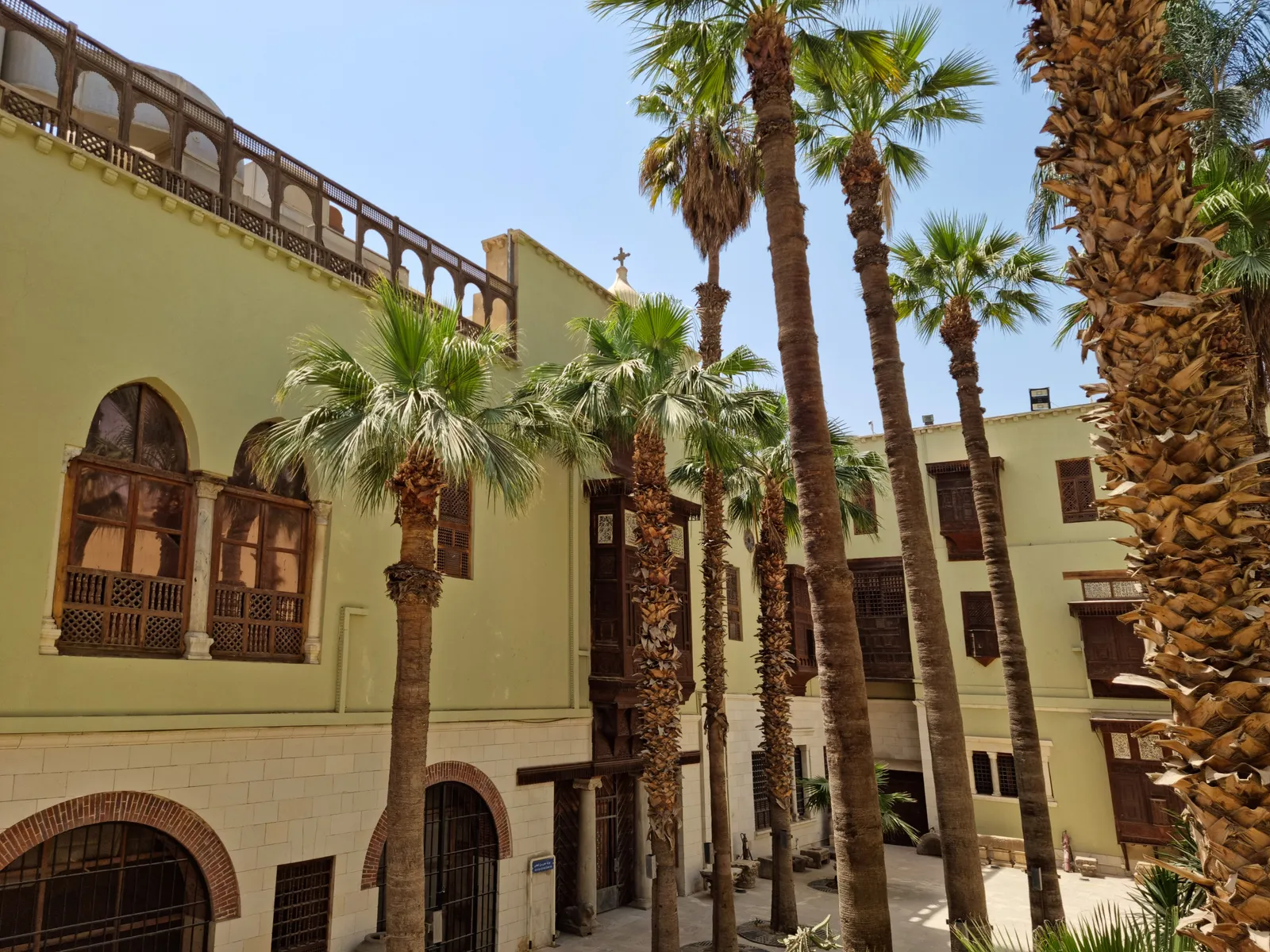 The Coptic Museum in Old Cairo seen from its garden, a pale two-storey building with carved wooden window screens and a first-floor arcade, date palms growing up in front of it