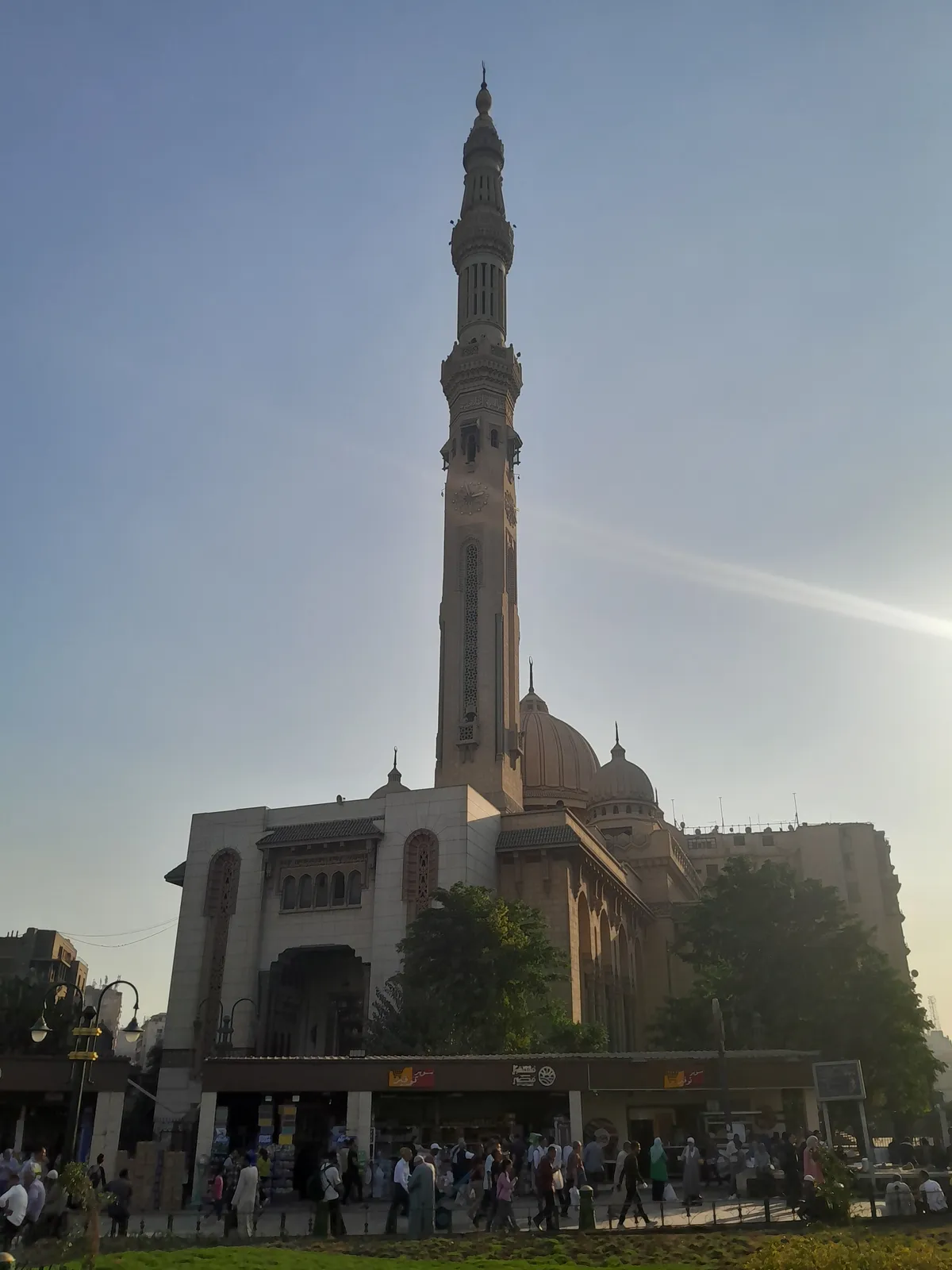 The single tall minaret of Al-Fath Mosque rising above Ramses Square in Cairo, with the mosque's domes and arcaded front below it and a crowd on the pavement outside the shopfronts