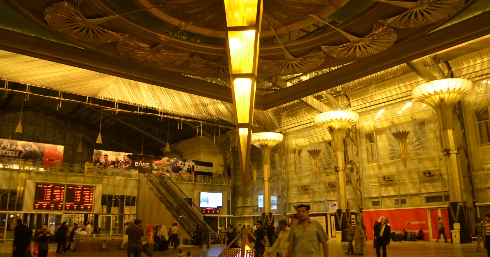 The main concourse of Ramses Station in Cairo under a fan-patterned ceiling, with tall lamp standards flaring open like capitals, departure boards along the far wall and passengers crossing the floor