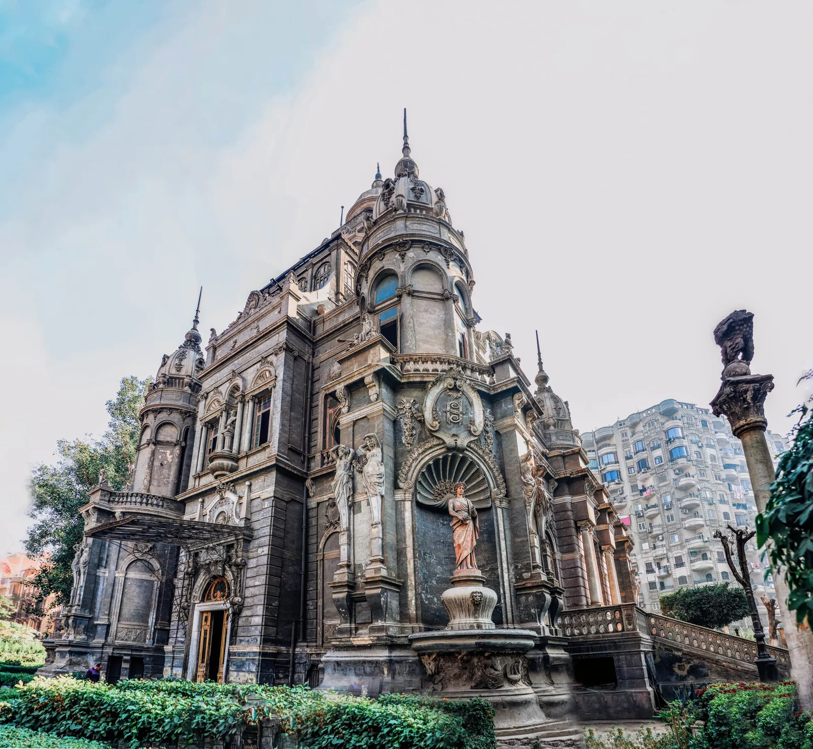 Sakakini Palace in Cairo seen from its garden, a grey stone mansion covered in rococo carving with turrets, statues in niches and a domed tower, modern apartment blocks rising behind it