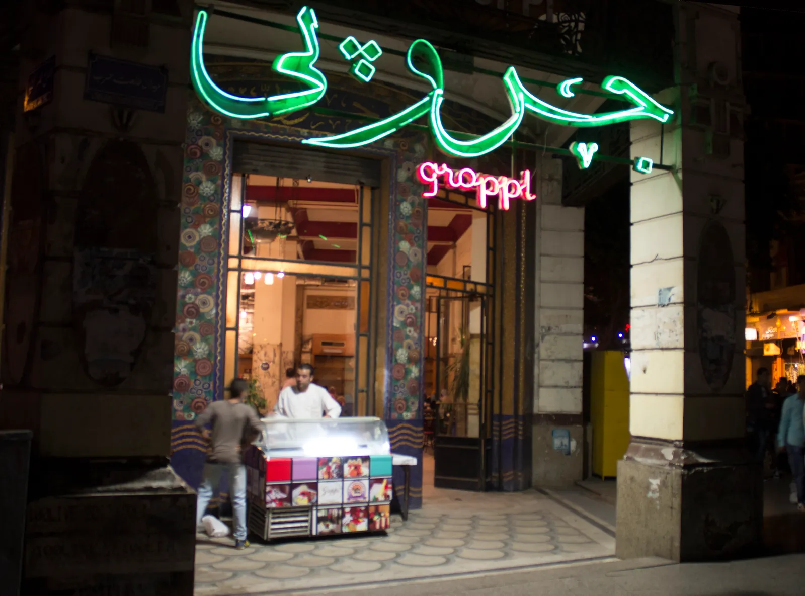 The entrance to Groppi on Talaat Harb Square in Cairo at night, an arched doorway framed by panels of coloured floral mosaic under a green and pink neon sign in Arabic and Latin lettering