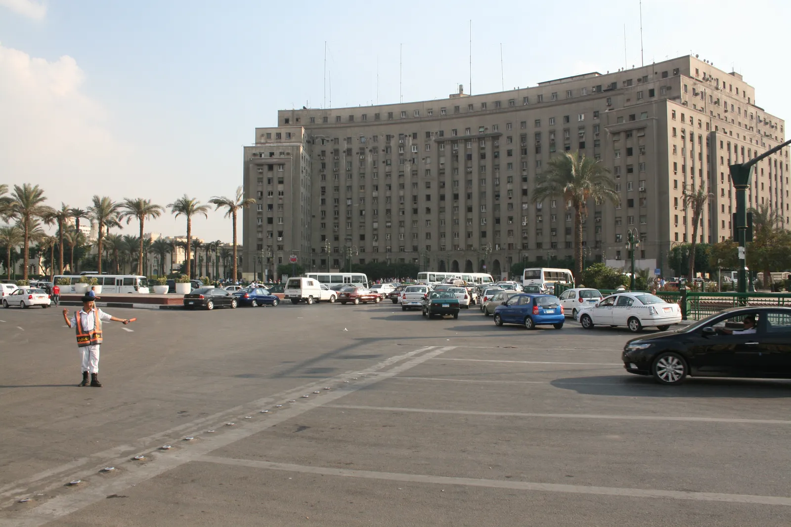 The Mogamma on Tahrir Square, a long curved government block of pale concrete rising fourteen storeys above a line of palms, with traffic crossing the open square in front of it
