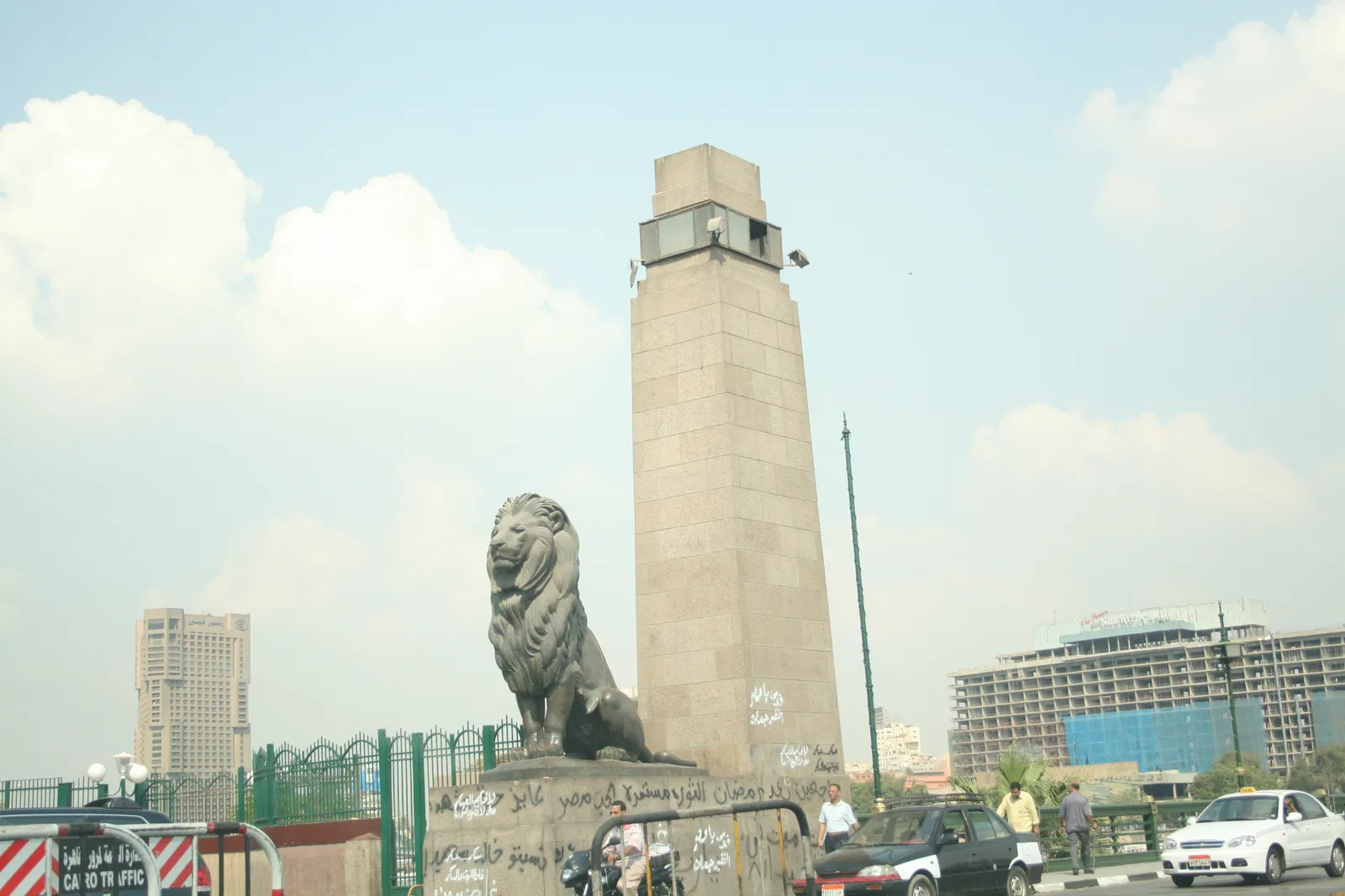 One of the bronze lions at the end of Qasr El Nil Bridge in Cairo, lying on a stone plinth beside the bridge's stone pylon with traffic and the far bank behind