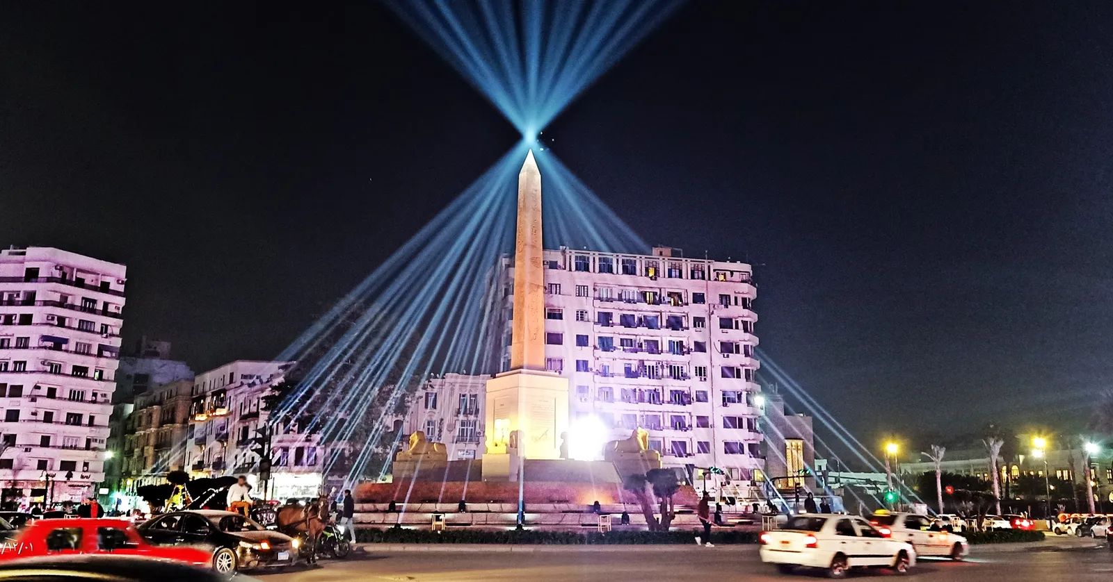 Tahrir Square in Cairo at night, a floodlit stone obelisk standing on a raised circular base in the middle of the traffic island with searchlight beams fanning out behind it and apartment blocks lit up around the square