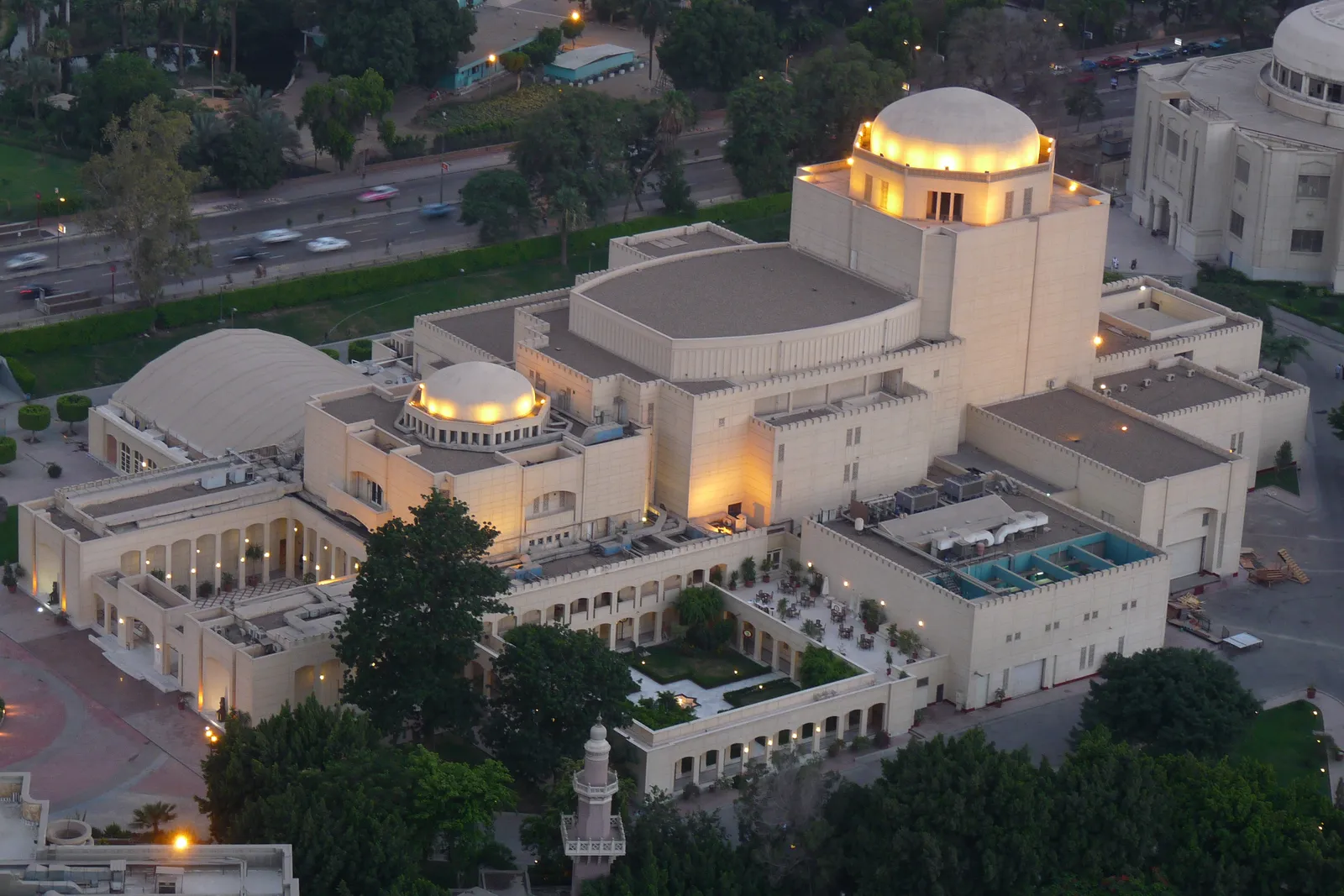The Cairo Opera House complex at dusk from above, a group of pale domed and colonnaded buildings lit along their roofs, set among lawns and trees with a road curving past