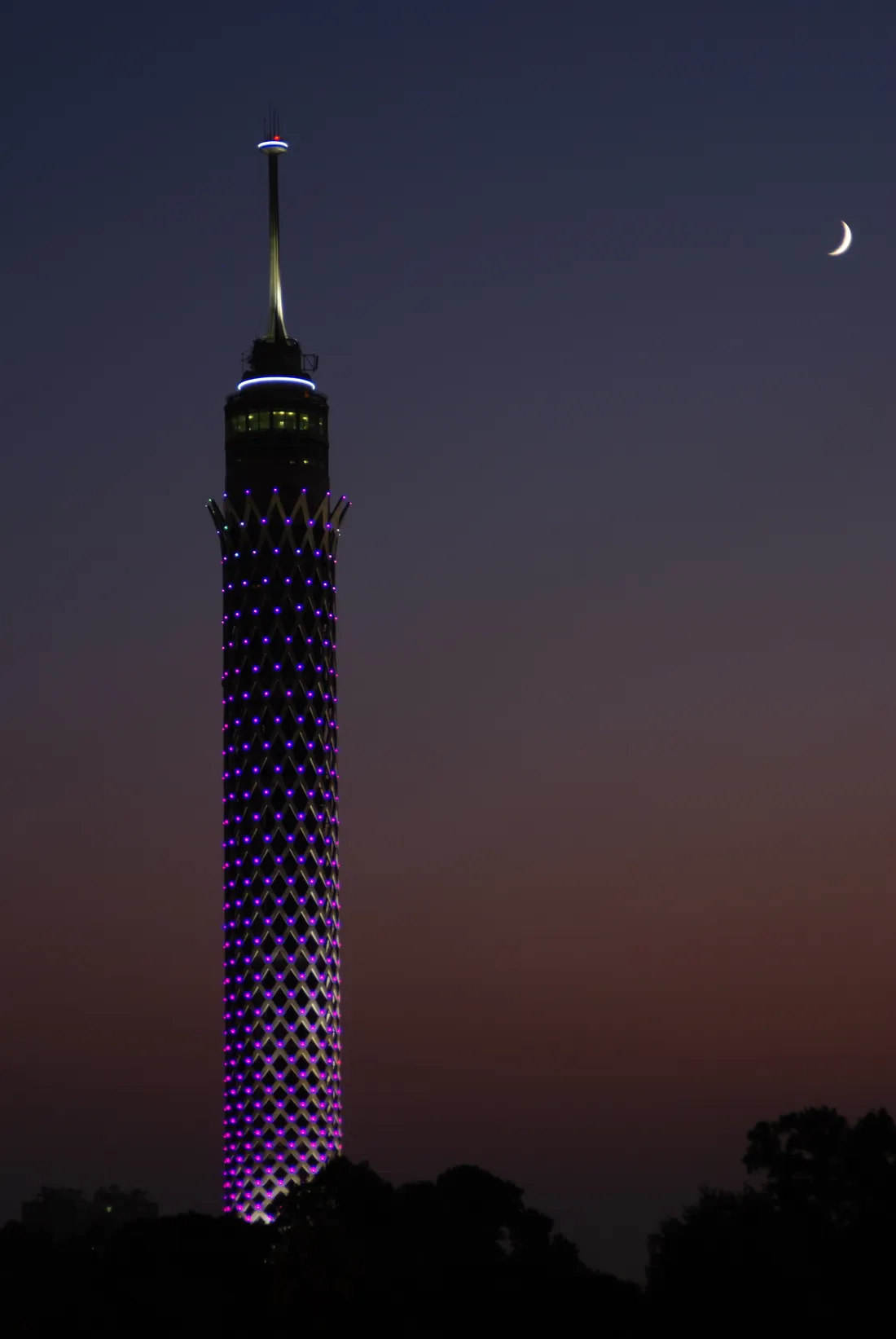 Cairo Tower at dusk, a tall concrete shaft wrapped in a diamond lattice lit from within, with the observation deck and mast at the top and a crescent moon in the darkening sky beside it