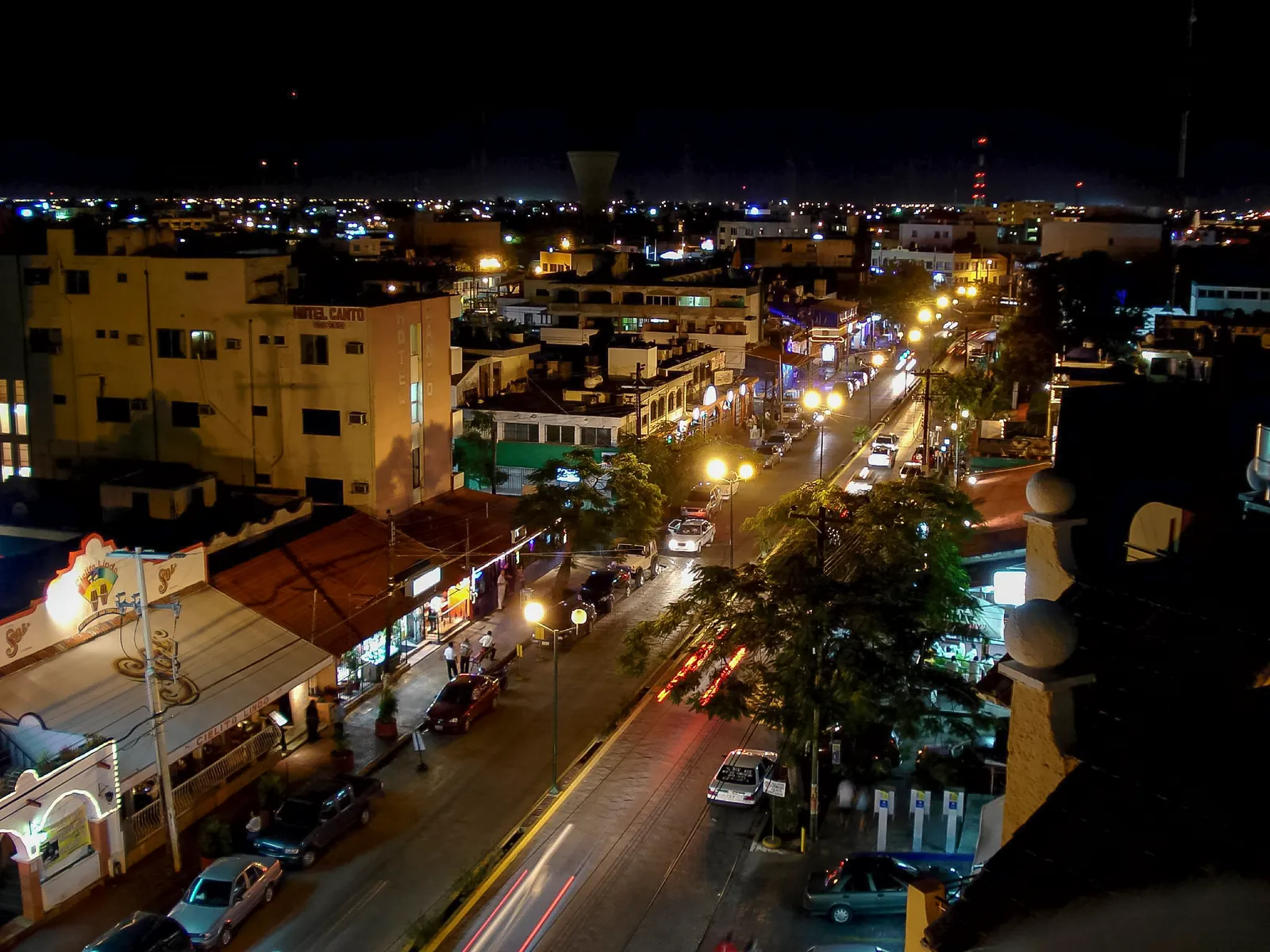 Avenida Yaxchilán in downtown Cancún seen from above at night, lit shopfronts and restaurant signs down both sides of a divided avenue with traffic and palm trees along the median