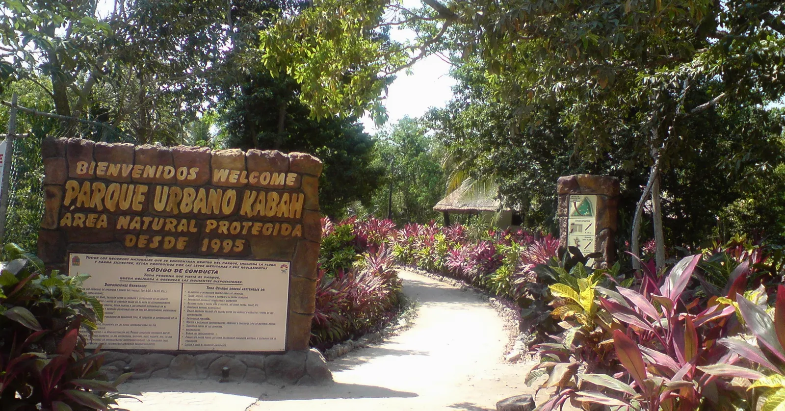 The entrance to Parque Urbano Kabah in Cancún, a carved welcome sign beside a pale path curving into dense green jungle with a thatched roof visible through the trees