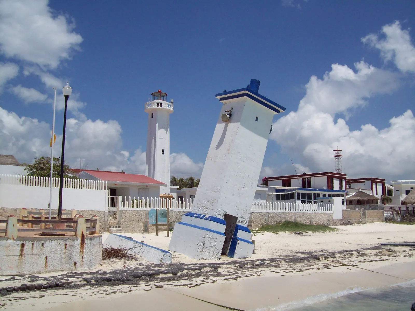 The old blue and white lighthouse at Puerto Morelos tilting steeply out of the sand beside the water, with the newer upright white lighthouse and low town buildings behind it