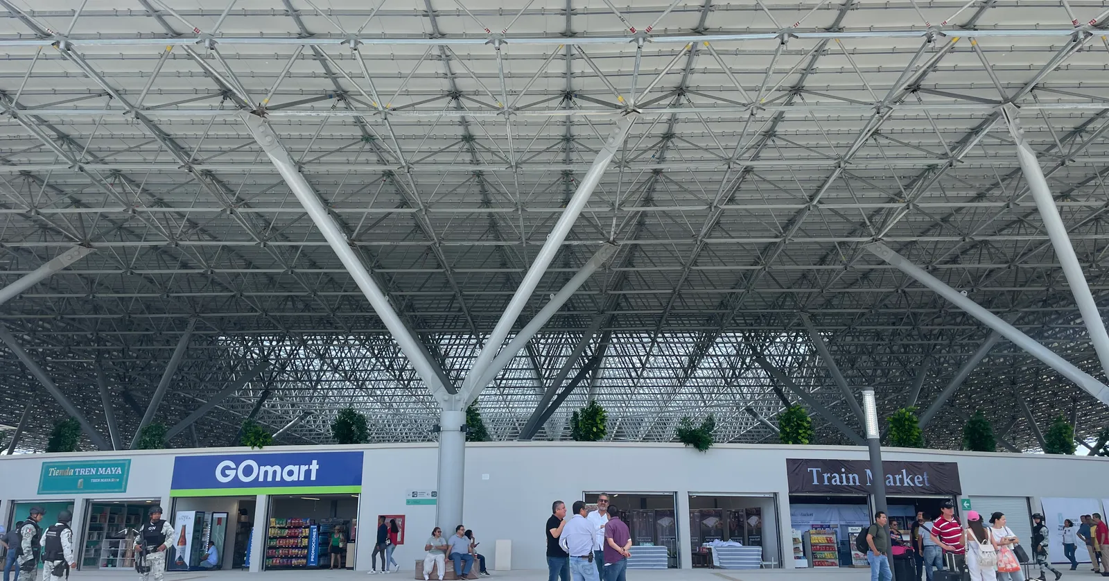 The Cancún Airport railway station on the Tren Maya line, a wide folded canopy carried on splayed white steel columns over an open concourse with shopfronts and travellers pulling luggage