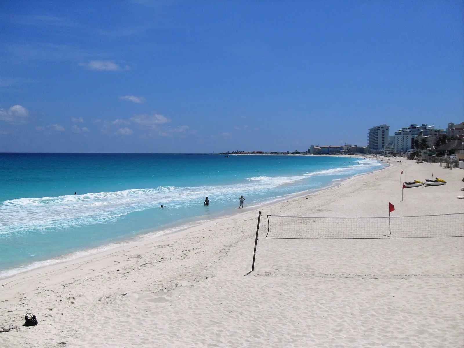 Playa Delfines in Cancún, a long stretch of white sand with lines of surf breaking onto it, a volleyball net and a red warning flag standing on the beach