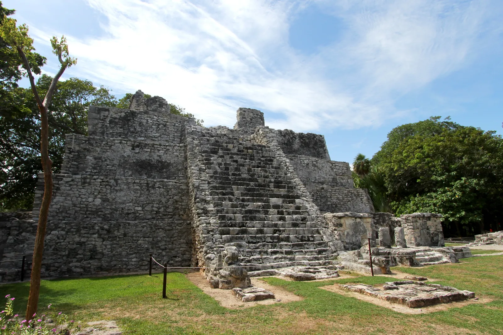 El Castillo at the El Meco archaeological site north of Cancún, a weathered stepped stone pyramid with a steep central staircase rising from mown grass under a blue sky