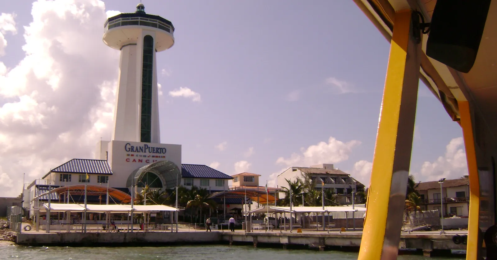 The Gran Puerto ferry terminal at Puerto Juárez in Cancún seen from the water, a tall white observation tower above low blue-roofed buildings along the quay