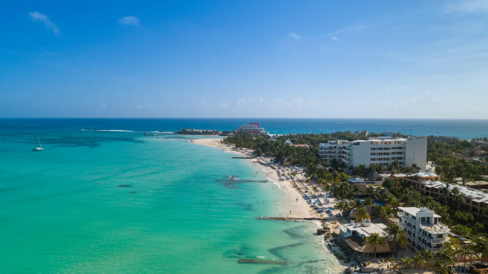 Playa Norte at the northern tip of Isla Mujeres from the air, a curve of white sand with pale turquoise shallows running far out from the shore and low buildings behind the beach