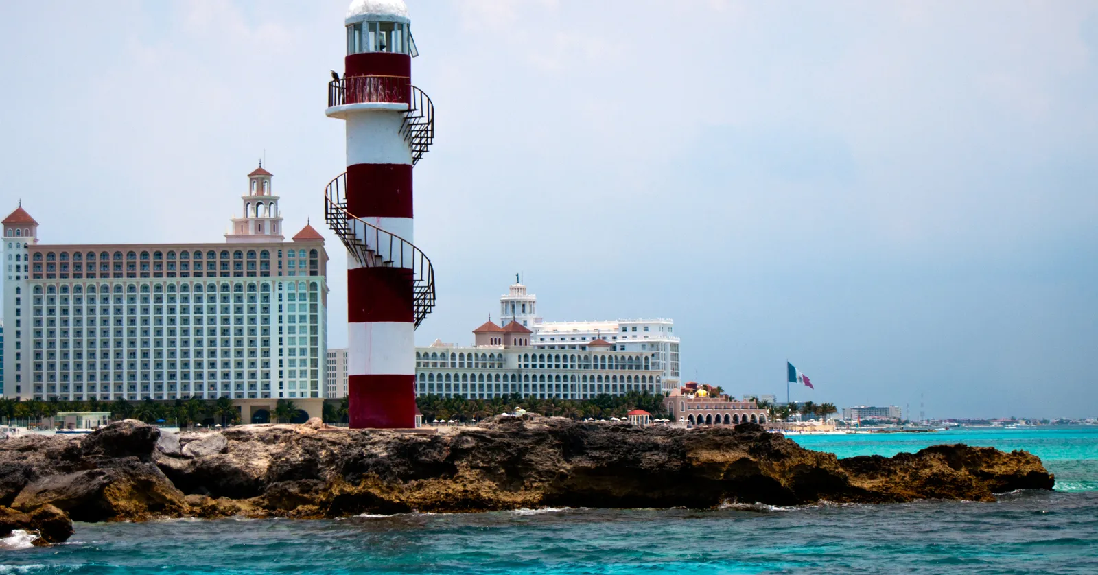 The red and white striped lighthouse at Punta Cancún standing on a low rocky shelf above turquoise water, with the pale hotel blocks of the Hotel Zone lined up behind it