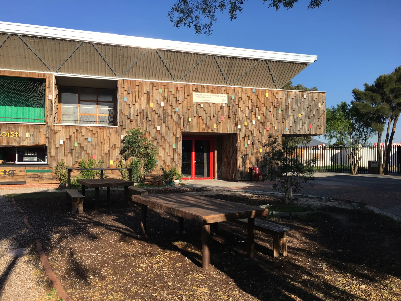 The Guga S'thebe theatre in Langa, Cape Town, its walls clad in strips of weathered timber dotted with small painted blocks, a red entrance door and wooden picnic tables on the ground in front