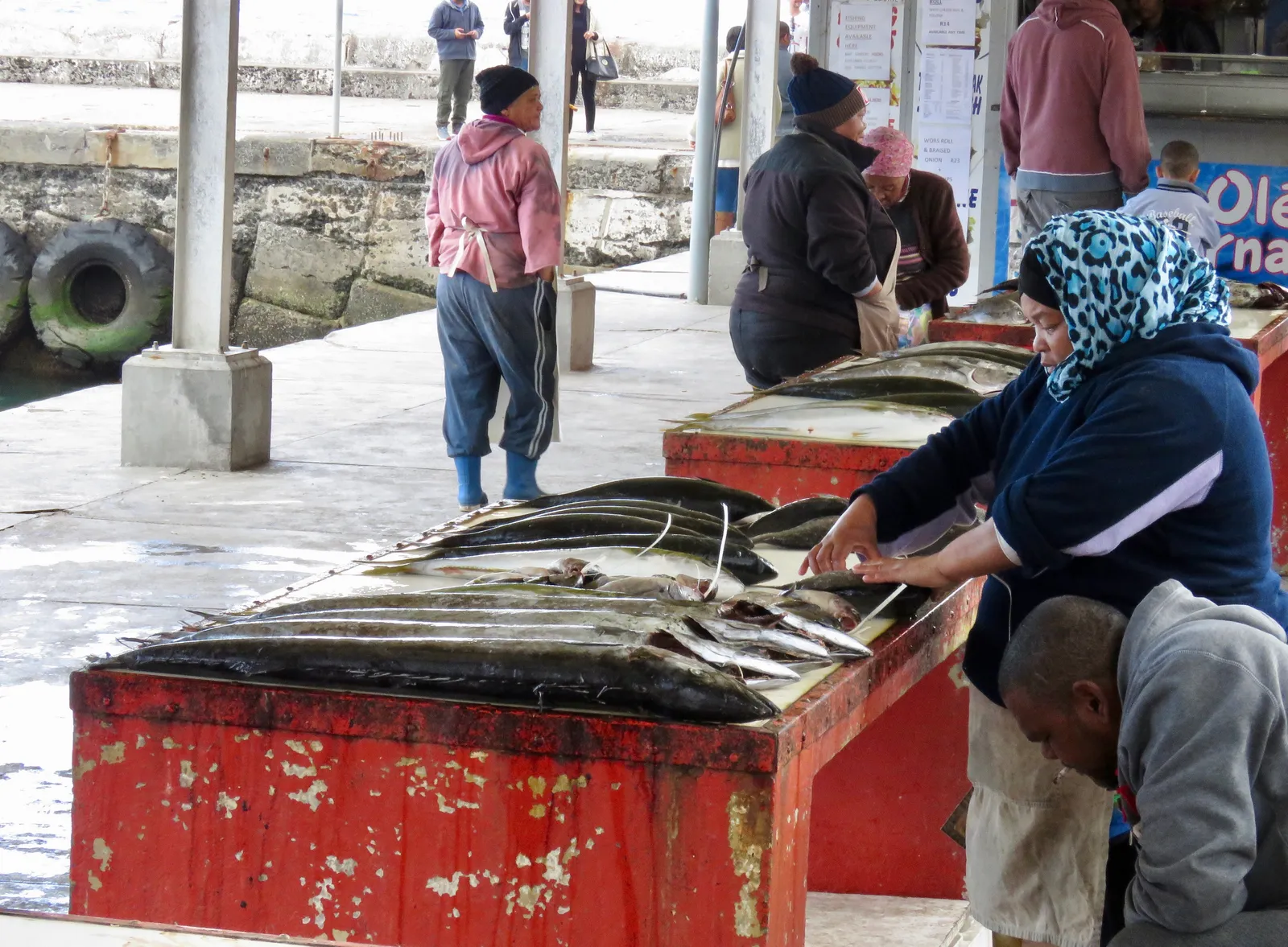 Fish laid out for sale on battered red tables on the quay at Kalk Bay harbour in Cape Town, a woman in a headscarf cleaning a fish while buyers in warm jackets wait