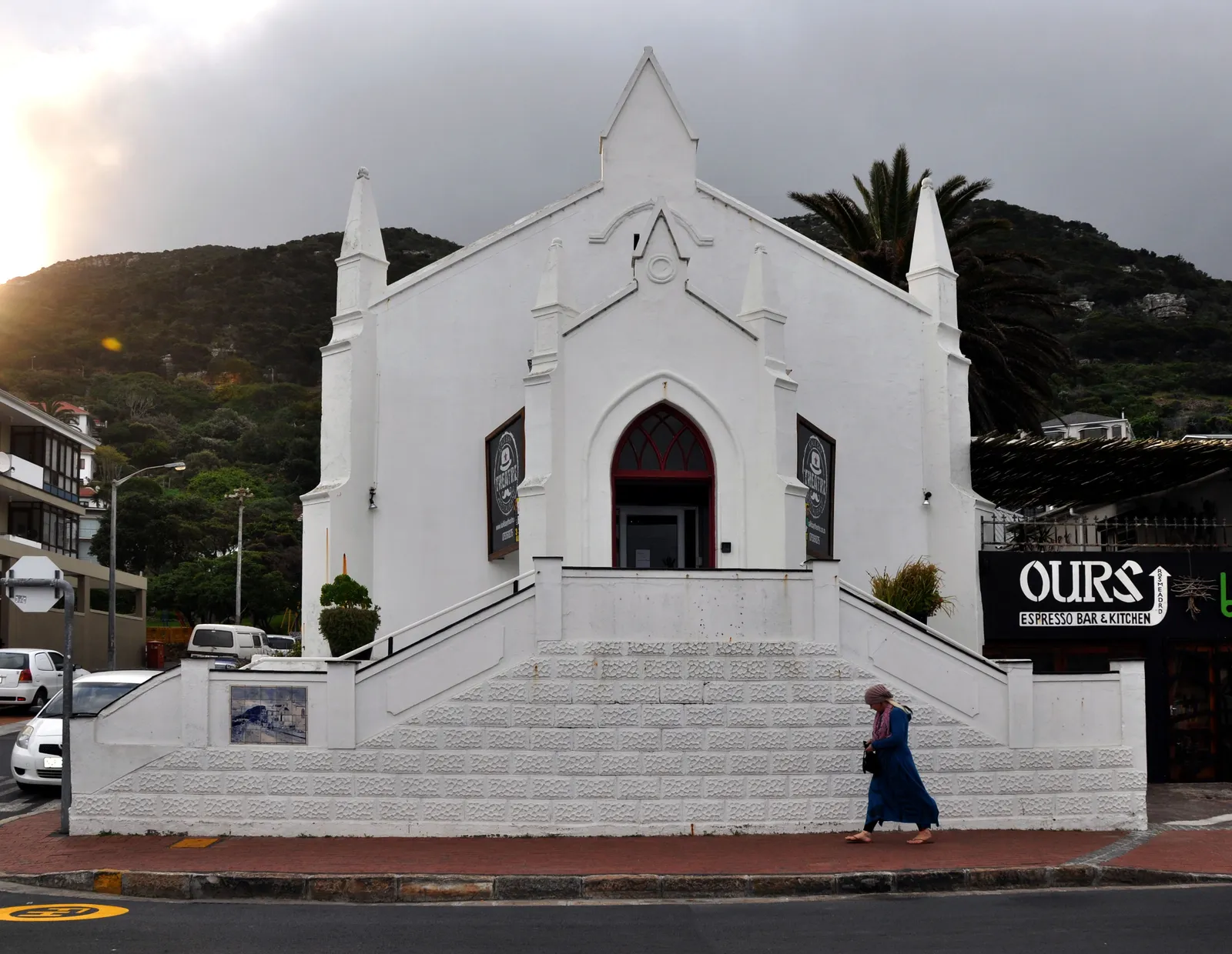 The Kalk Bay Theatre on Main Road in Cape Town, a white neo-Gothic former church with pointed pinnacles and a dark red arched door at the top of a wide flight of steps, under a cloudy sky with the mountain slope behind
