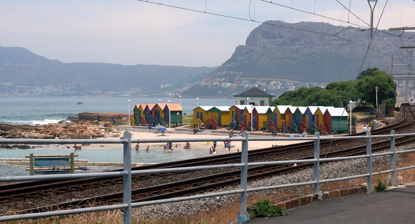 A row of bathing boxes painted in bright stripes of red, yellow, blue and green on the beach at St James in Cape Town, beside a tidal pool, seen across the railway tracks with False Bay and the mountains behind