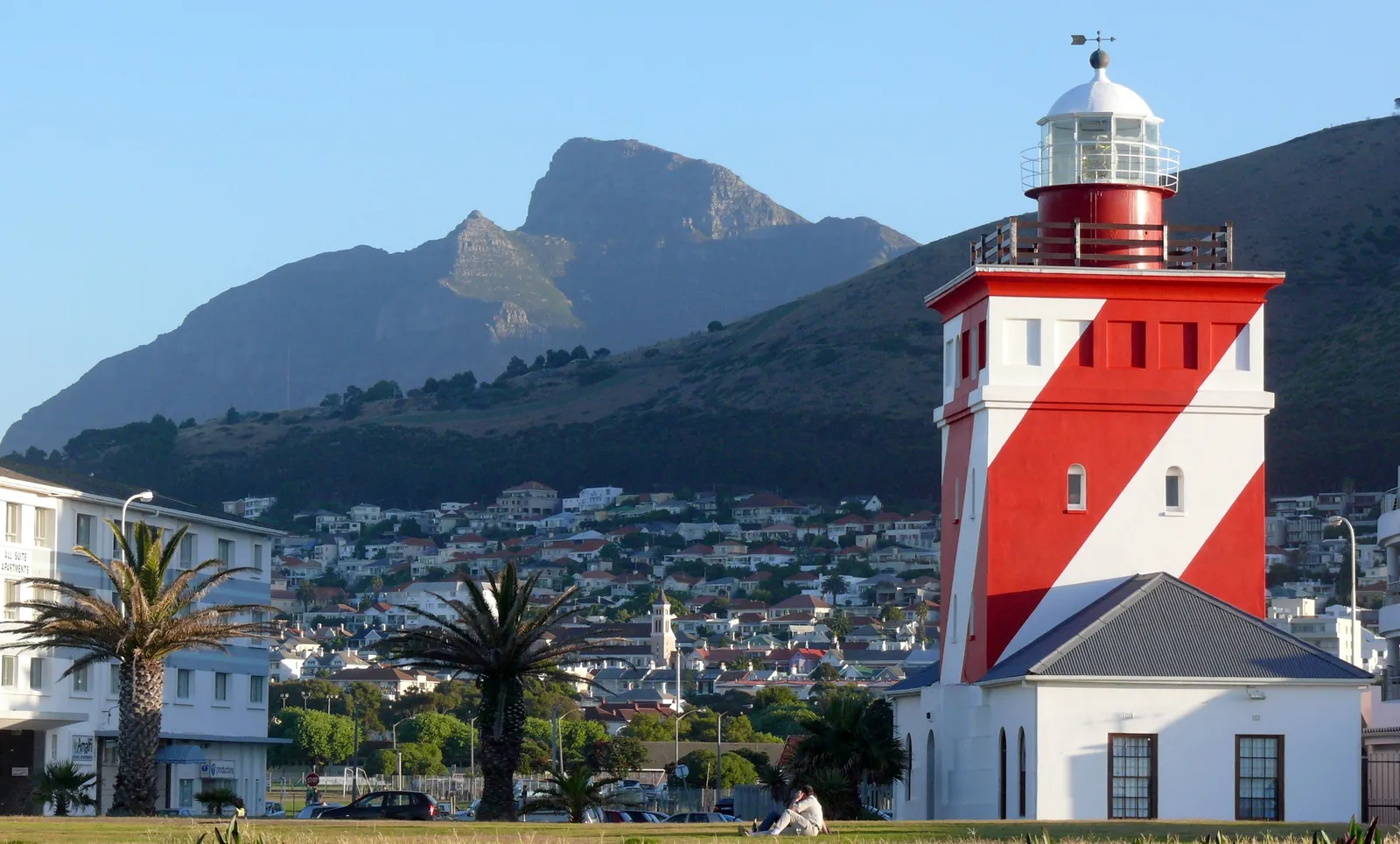 The Green Point Lighthouse at Mouille Point in Cape Town, a square tower painted with bold red and white diagonal stripes and topped by a red lantern gallery, with palm trees, houses on Signal Hill and the cliffs of Table Mountain behind