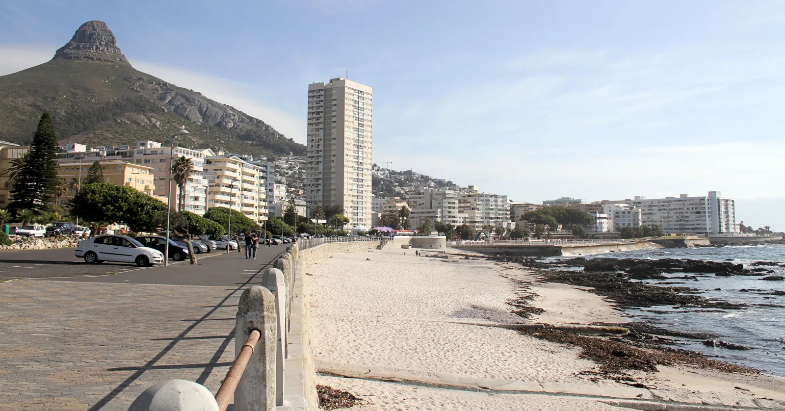 The Sea Point Promenade in Cape Town, a wide paved walkway with a low concrete railing above a white sand beach and rocky shore, lined with apartment blocks and palm trees, with the pointed peak of Lion's Head rising behind