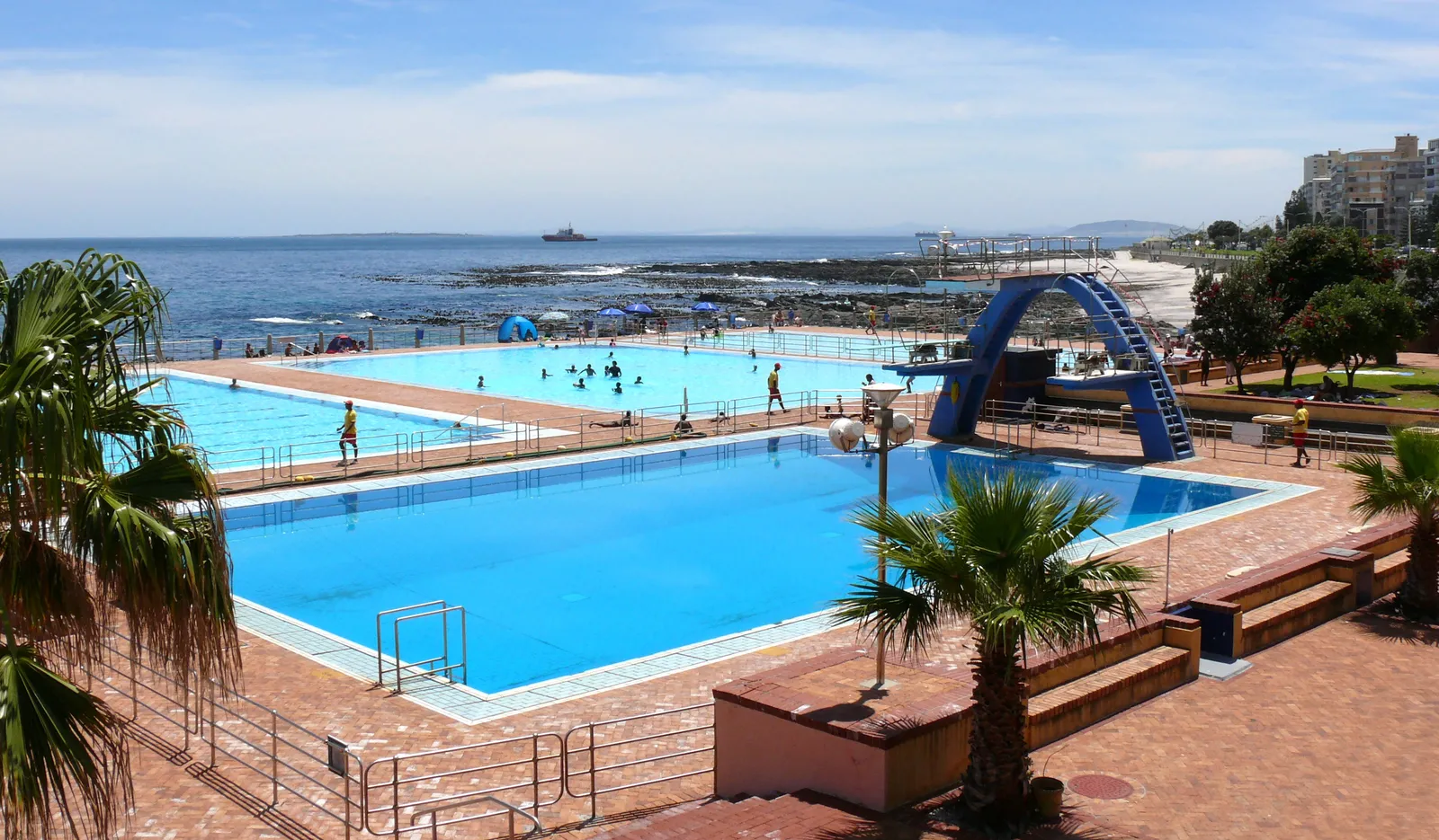 The open-air pools of the Sea Point Pavilion in Cape Town, a large blue swimming pool and a blue diving tower in the foreground, more pools beyond and the Atlantic Ocean behind a low sea wall