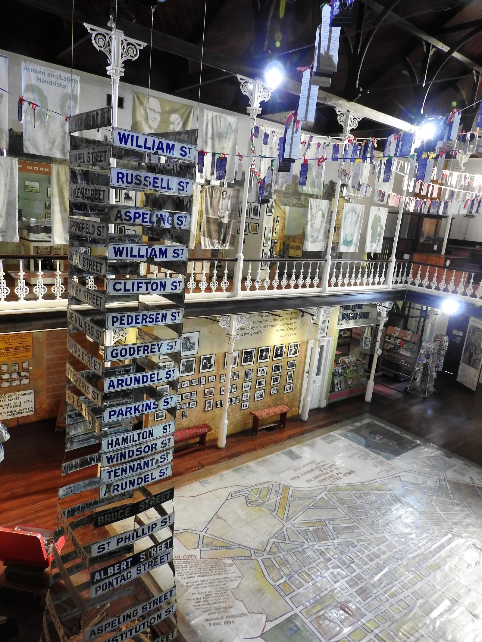 Inside the District Six Museum in Cape Town, a tall stack of old blue and white street signs beside a large painted map of the district on the floor, under a gallery hung with banners and photographs