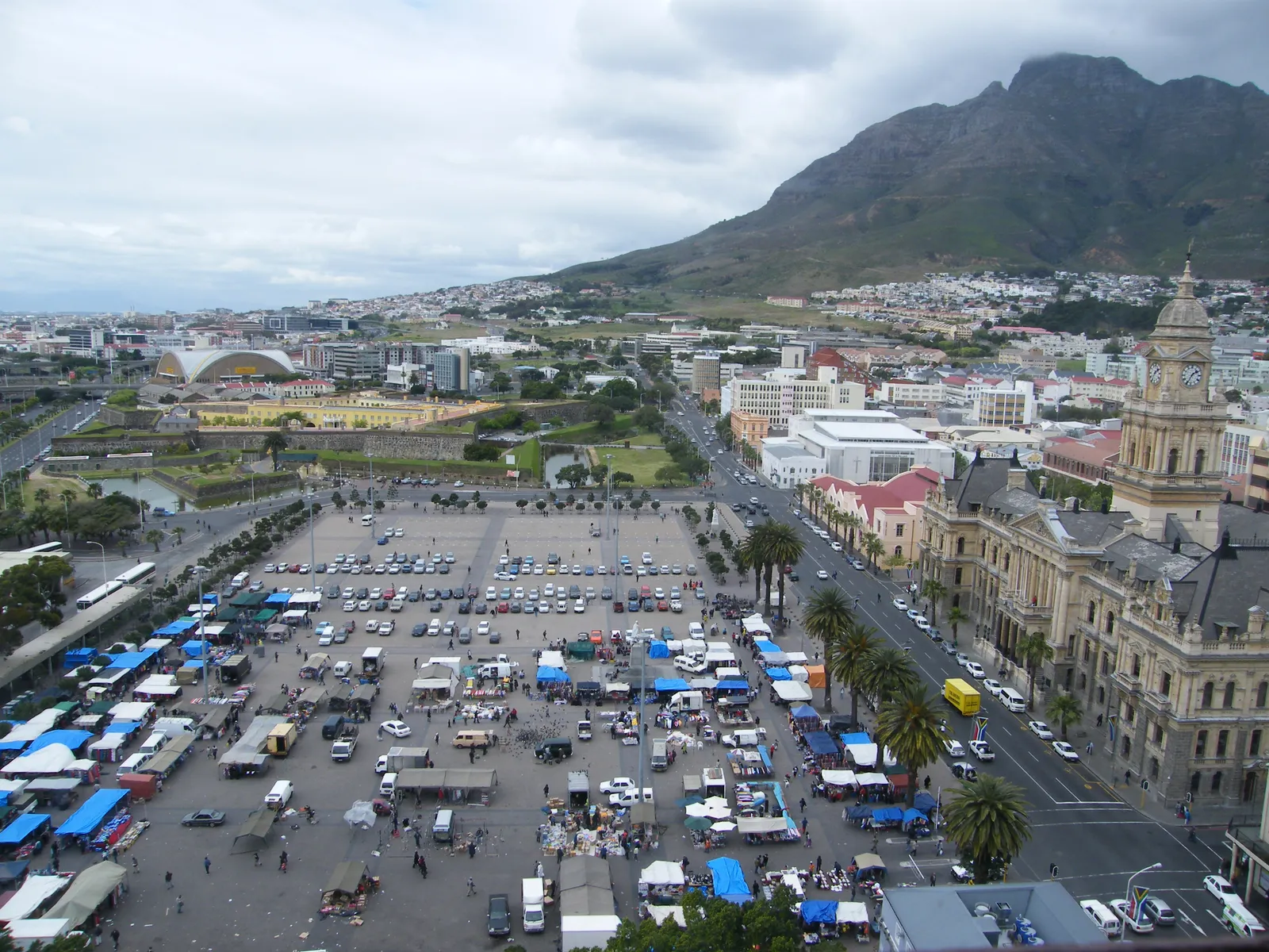 Cape Town's Grand Parade seen from above, market stalls under blue and white awnings among parked cars, with the sandstone City Hall and its clock tower on the right, the Castle of Good Hope's walls and moat behind, and Devil's Peak rising over the city