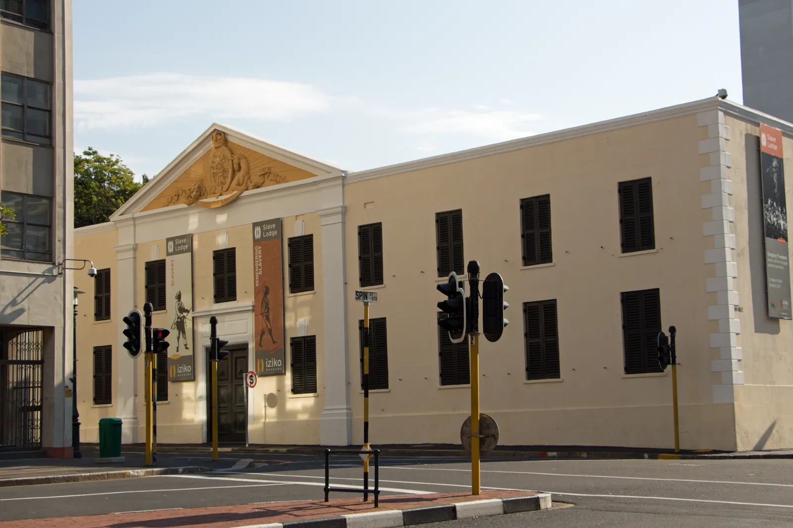 The Slave Lodge in Cape Town, a long cream two-storey building with dark shuttered windows and a carved relief in its pediment, seen from a street corner with traffic lights