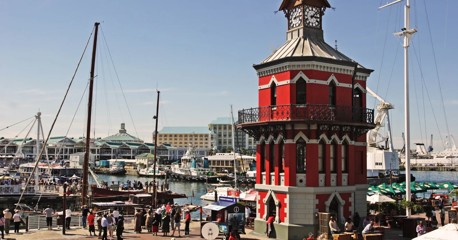 The red and white Victorian Gothic Clock Tower at Cape Town's V&A Waterfront, with an iron balcony and a clock face at the top, beside a harbour basin full of tugs and sailing masts and a crowd on the quayside
