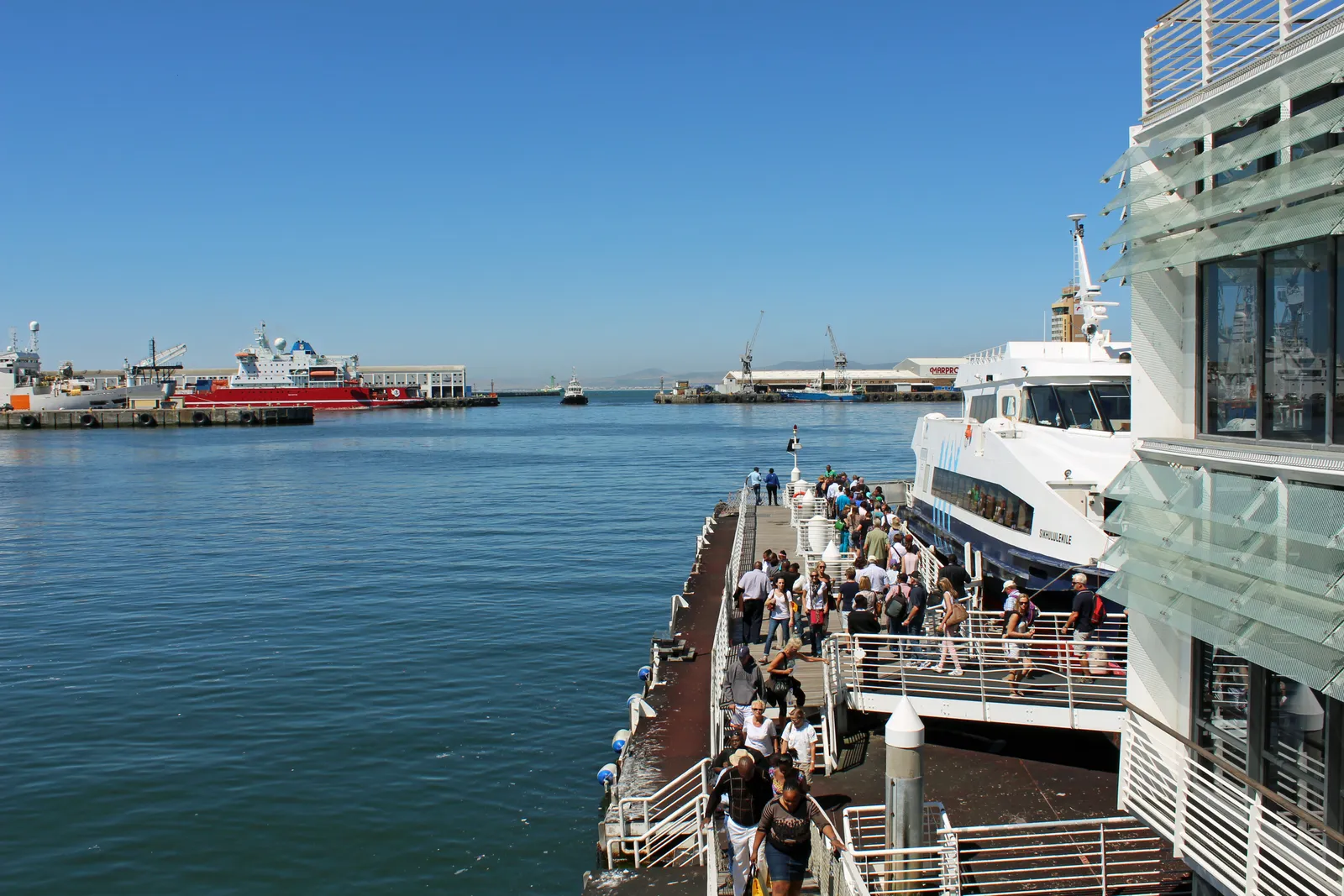 Passengers filing down a covered gangway onto a white ferry moored beside the Nelson Mandela Gateway at Cape Town's V&A Waterfront, with the harbour and docked ships beyond under a clear sky