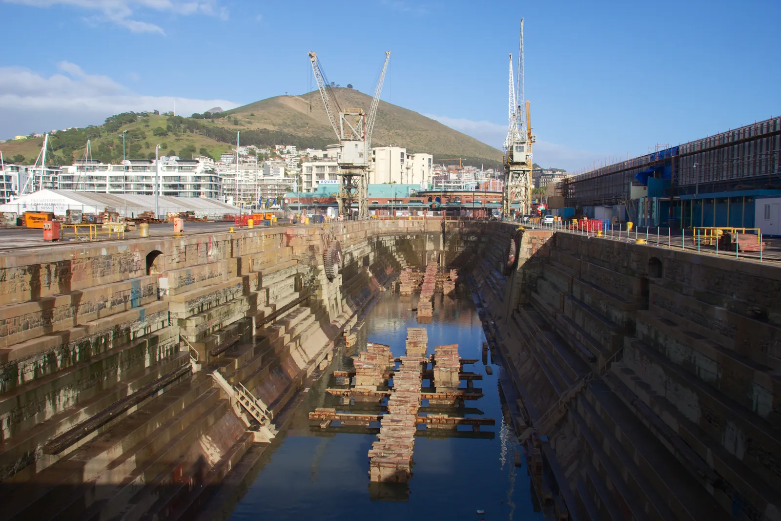 The drained Robinson Dry Dock at Cape Town's V&A Waterfront, stepped stone walls running down to a strip of water and a line of wooden keel blocks, with cranes and Signal Hill behind