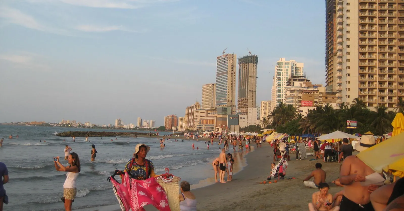 Bocagrande beach in Cartagena at golden hour, with beachgoers, a vendor carrying goods, and the neighborhood's high-rise towers lining the shore