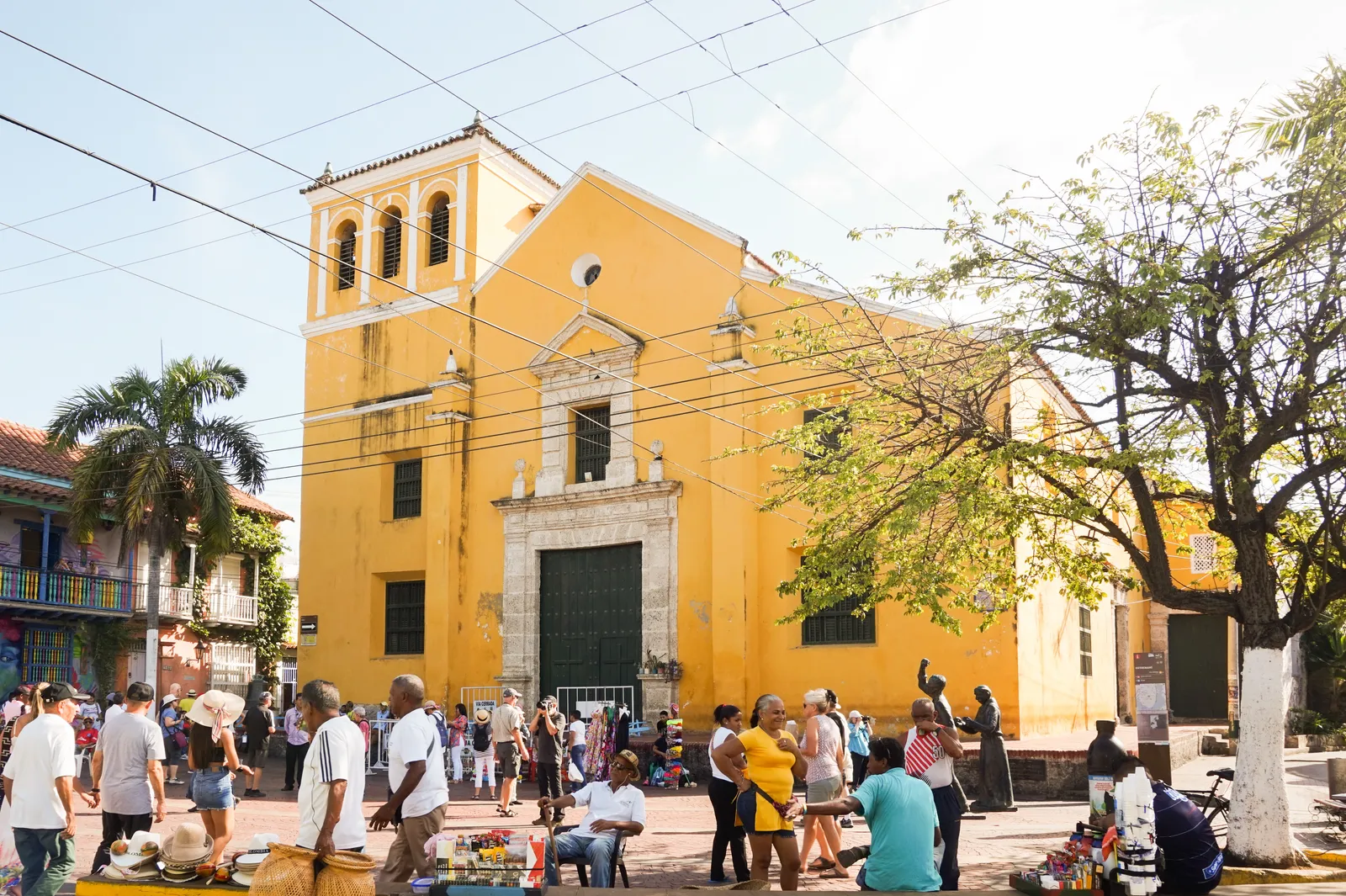 The yellow colonial facade and bell tower of the Iglesia de la Santisima Trinidad on Plaza de la Trinidad in Cartagena, with vendors and visitors gathered outside