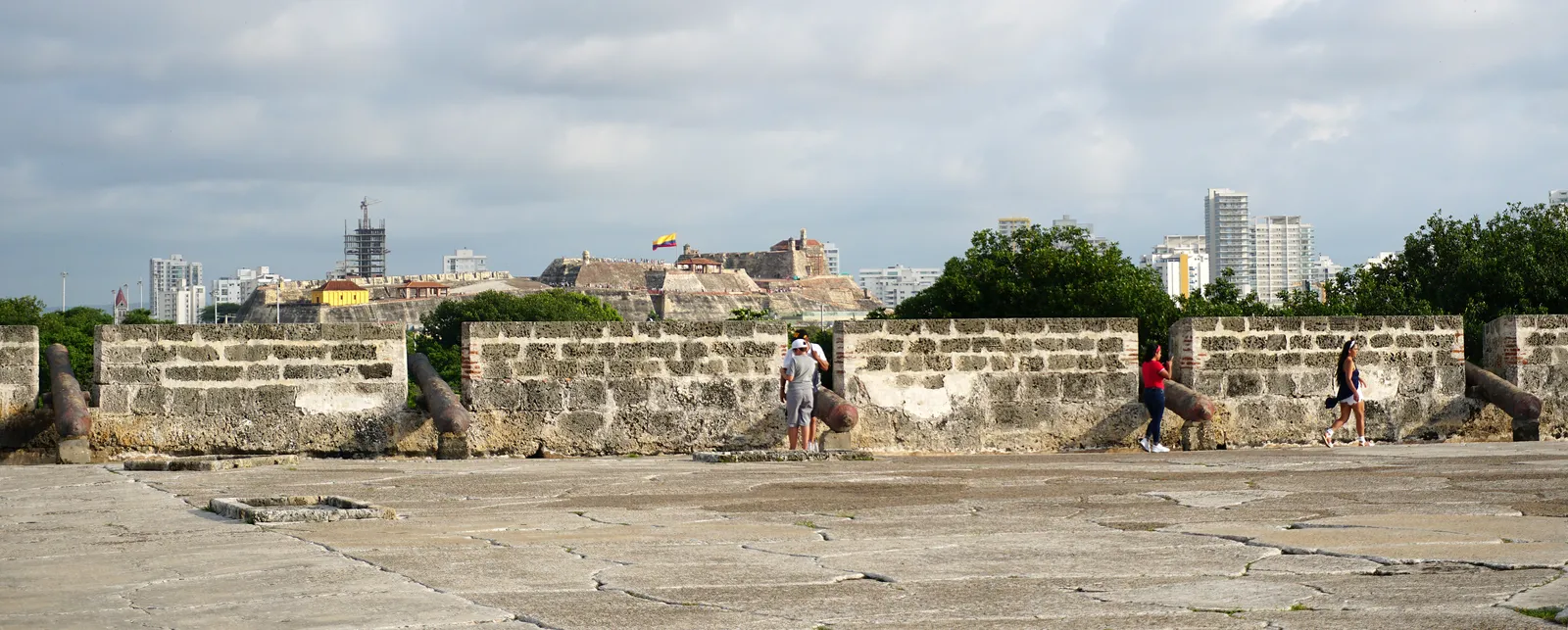 Old cannons mounted along a stone battery wall at Castillo San Felipe de Barajas, with visitors walking past and Cartagena's skyline visible behind the wall