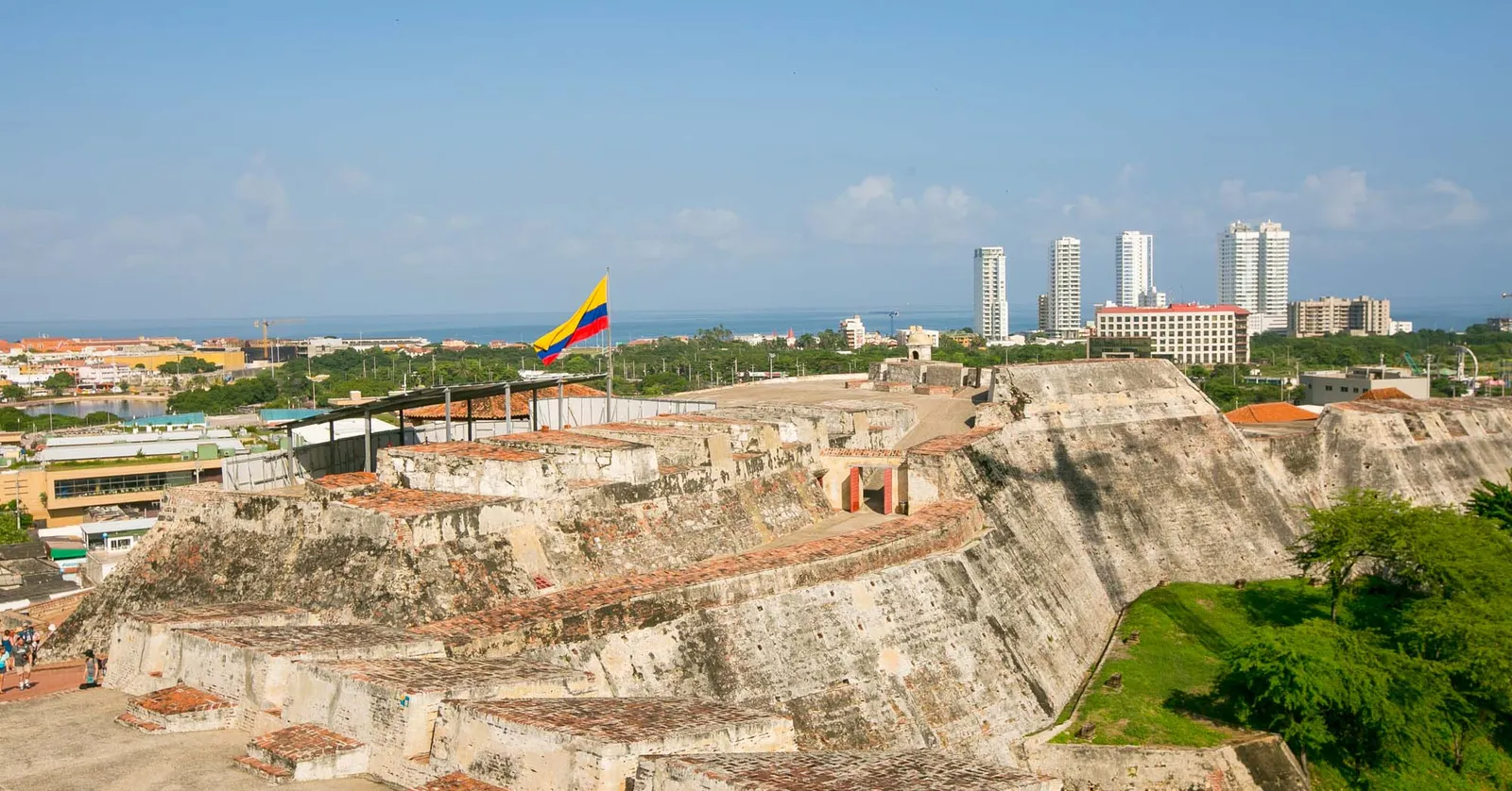The terraced stone walls of Castillo San Felipe de Barajas with a Colombian flag flying above them and the high-rise towers of Bocagrande visible in the distance