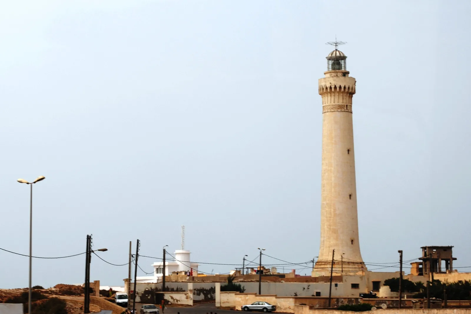 The El Hank lighthouse in Casablanca, a tapering pale stone tower with a balcony and lantern on top, standing above low flat-roofed houses with street lights and power lines in front of it