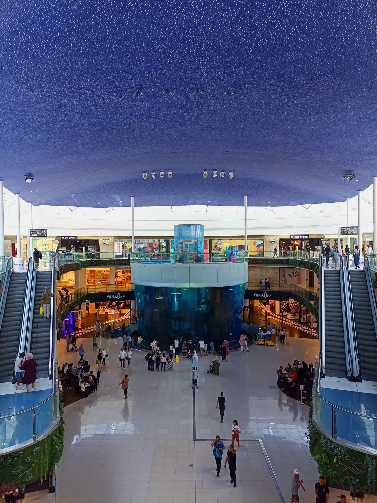 The central atrium of Morocco Mall in Casablanca, a tall cylindrical glass aquarium standing in the middle of the floor with people gathered around its base, escalators rising on both sides and a dark blue vaulted ceiling above