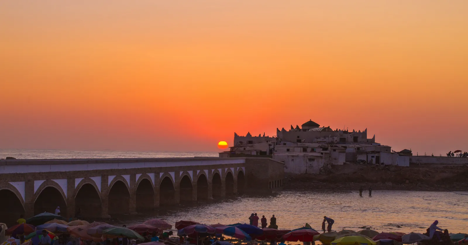 The shrine of Sidi Abderrahman in Casablanca at sunset, whitewashed houses and a domed tomb covering a rock just offshore, an arched footbridge running out to it and beach umbrellas packed along the sand in the foreground