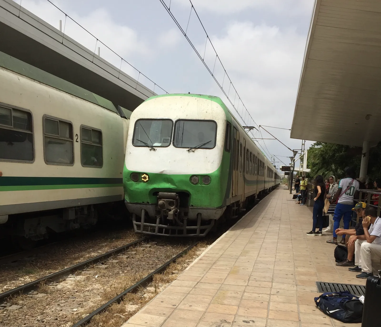 A green and cream commuter train standing at the platform at Casa-Oasis station in Casablanca, a second train alongside it, passengers waiting on a tiled platform under a concrete canopy