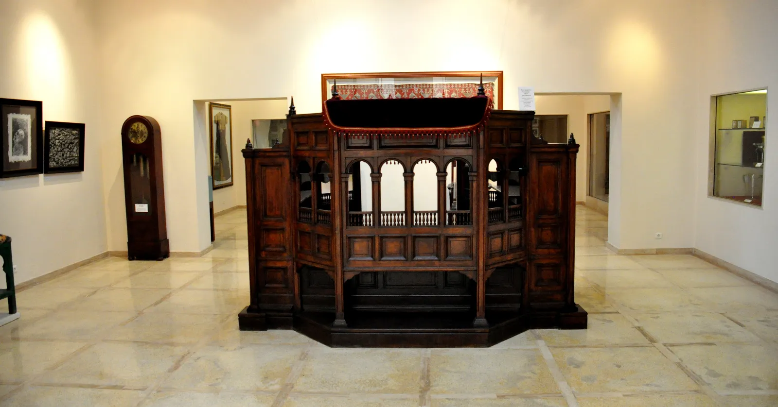 An exhibition hall at the Museum of Moroccan Judaism in Casablanca, a carved dark-wood synagogue reading platform standing alone in the middle of a white gallery, with a longcase clock and framed works along the walls
