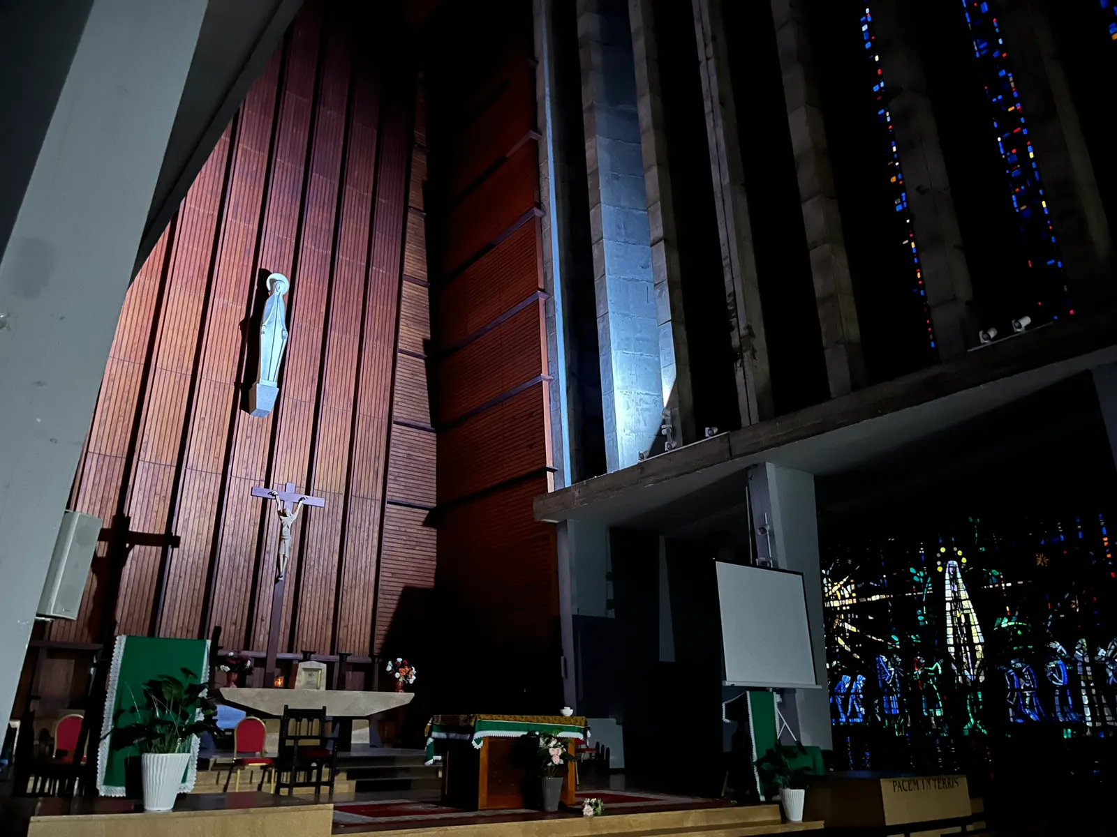Inside Notre-Dame de Lourdes church in Casablanca, a tall panelled timber wall behind the altar with a small statue of the Virgin on it, and floor-to-ceiling bands of deep blue and red stained glass between concrete ribs to the right