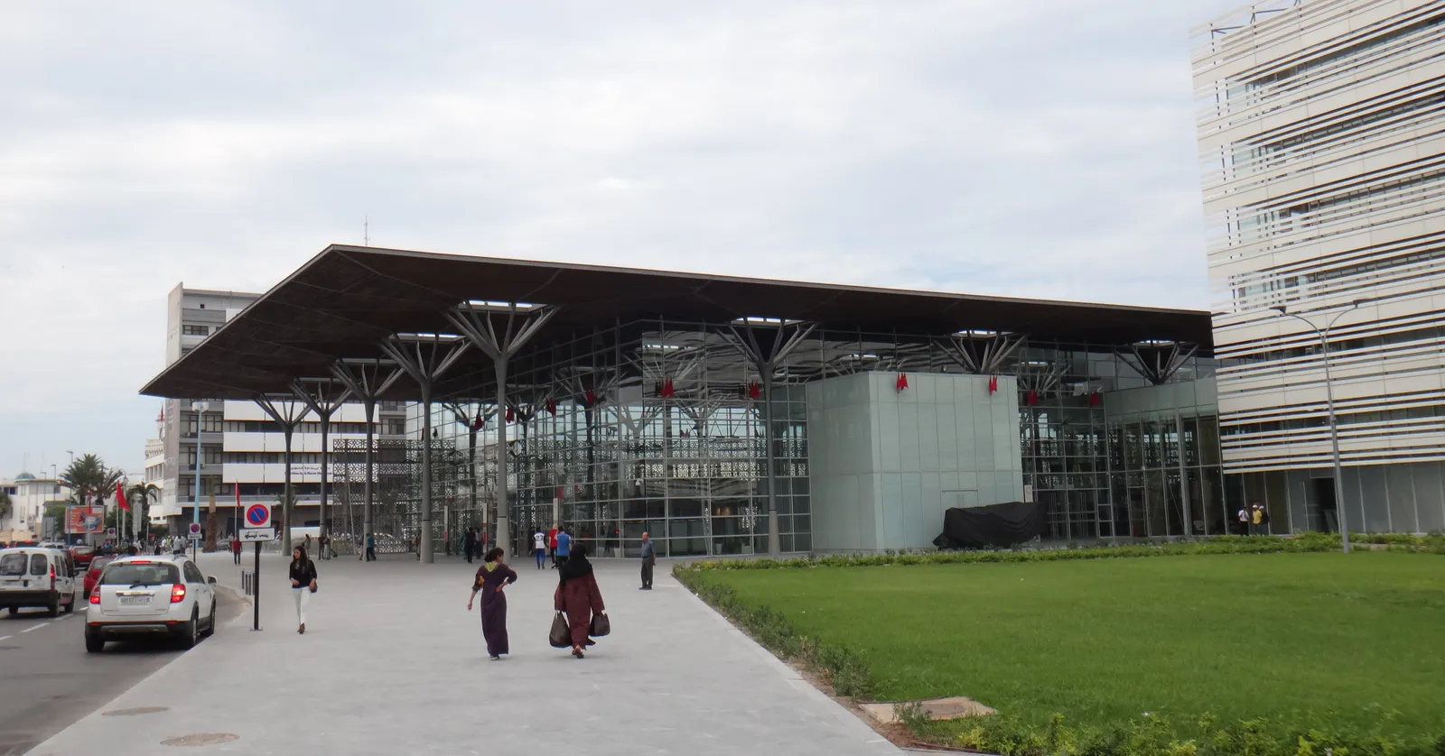 The Casa-Port railway station in Casablanca seen from the street, a broad flat roof canopy carried on slim tapering columns above a fully glazed hall, with people walking along the pavement in front of it